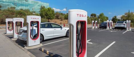 A parking area with multiple Tesla electric vehicle charging stations, a white car parked, and a modern building in the background.