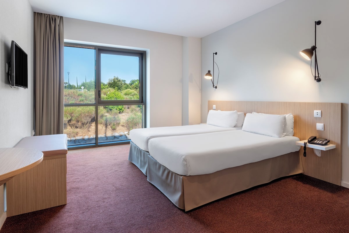 Habitación de hotel con una cama grande, un escritorio, un televisor y una ventana con vistas a la vegetación.