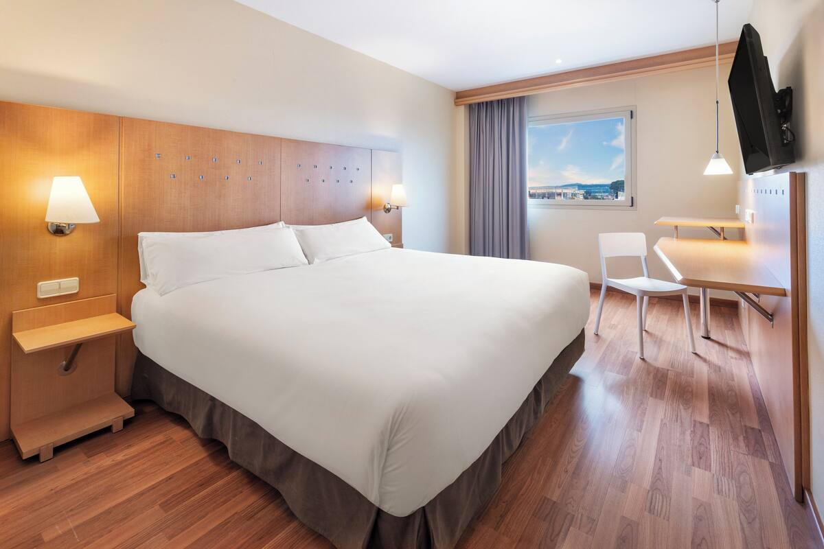 Habitación de hotel con una cama grande, mesitas de noche, escritorio, silla, televisor y ventana con vistas.