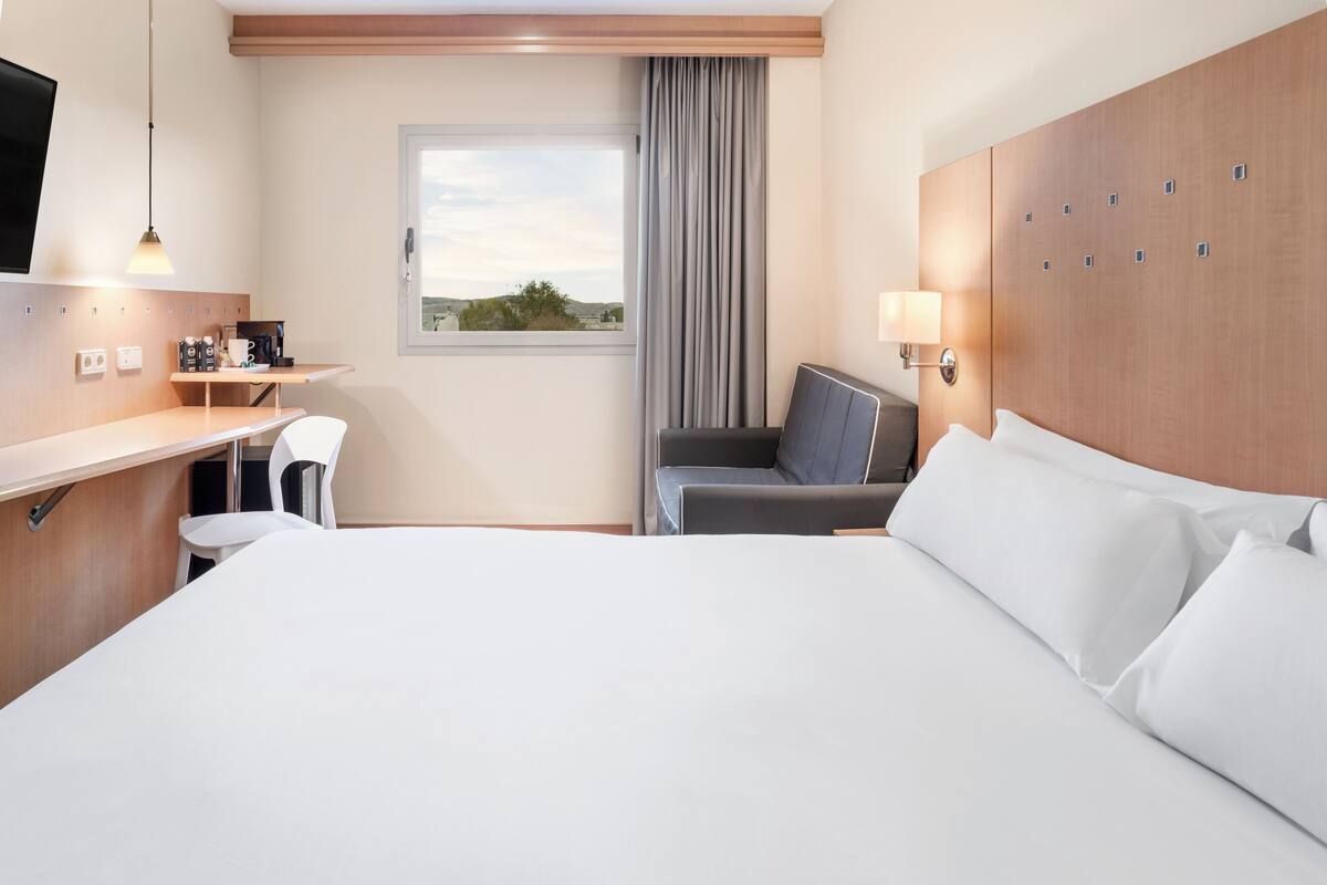Habitación de hotel con una cama grande, un escritorio con silla, un televisor, una ventana con vistas y una lámpara.