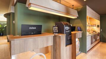 A reception desk with a digital kiosk, a coffee machine, and a breakfast bar with various food items.