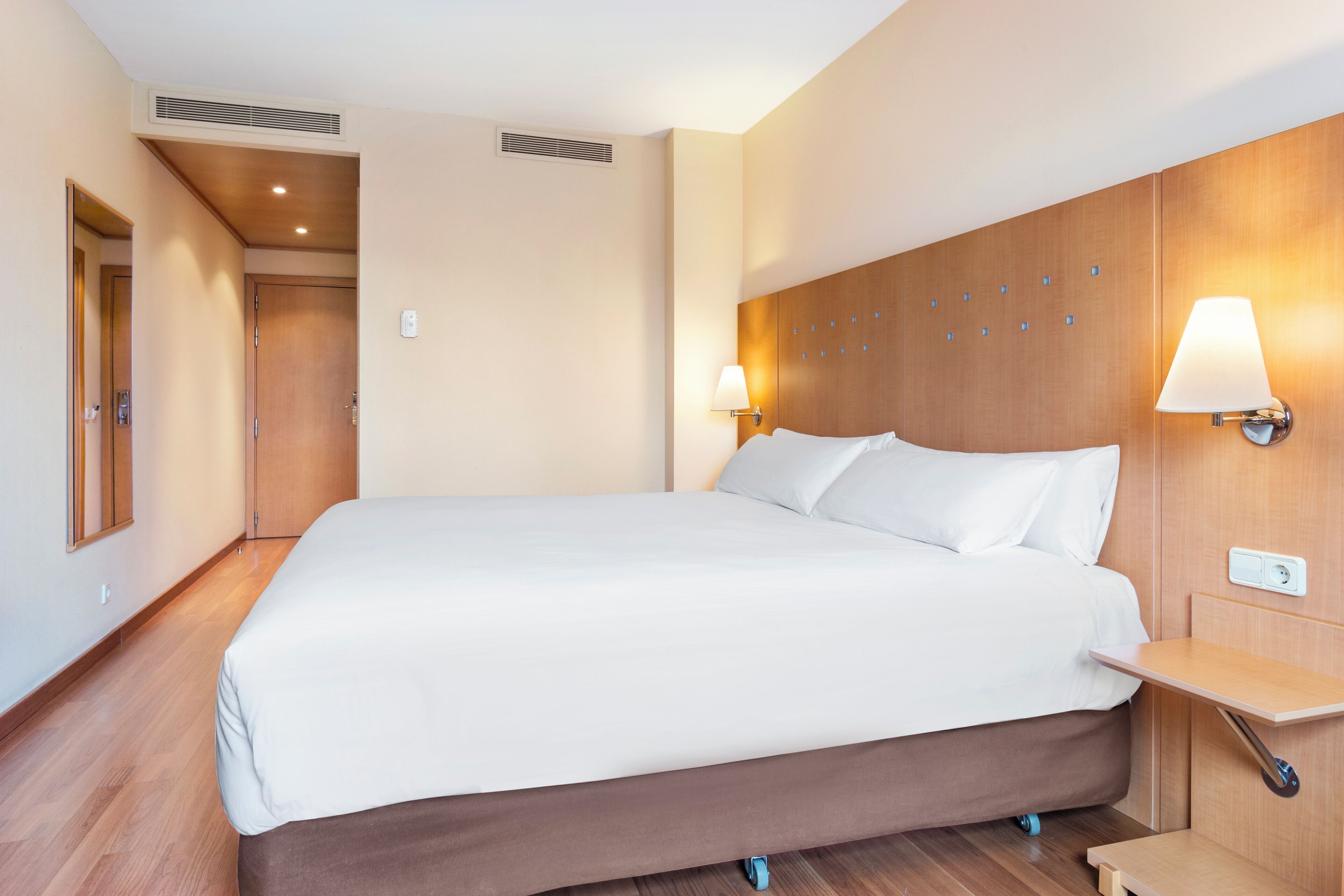 A hotel room with a large bed, two bedside lamps, a wooden headboard, and a small wooden side table.