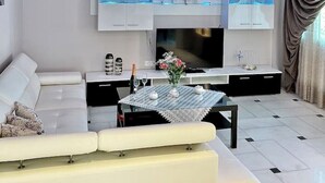 A modern living room with a white sofa, a coffee table, a flat-screen TV, and built-in shelving with decorative items. - Marble Luxury Villa in Neapolis (Αγιος Νικολαος)