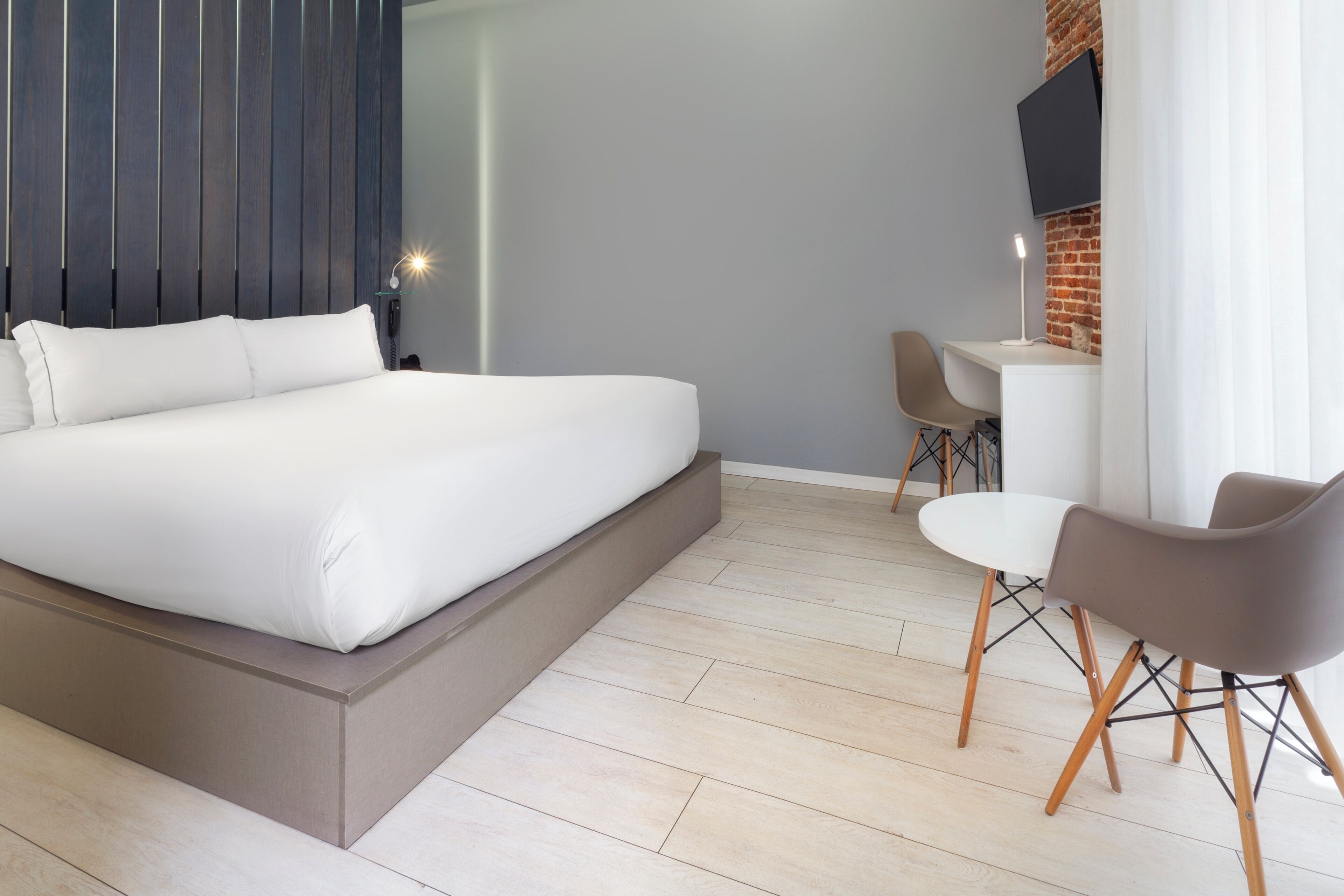 Un dormitorio moderno con una cama grande, un escritorio, dos sillas y un televisor fijado a una pared de ladrillo.