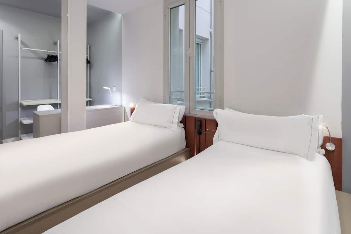 A hotel room with two single beds, a bedside table with a lamp, a mirror, a window with a view of the outside, and a built-in wardrobe with a hanging rack.