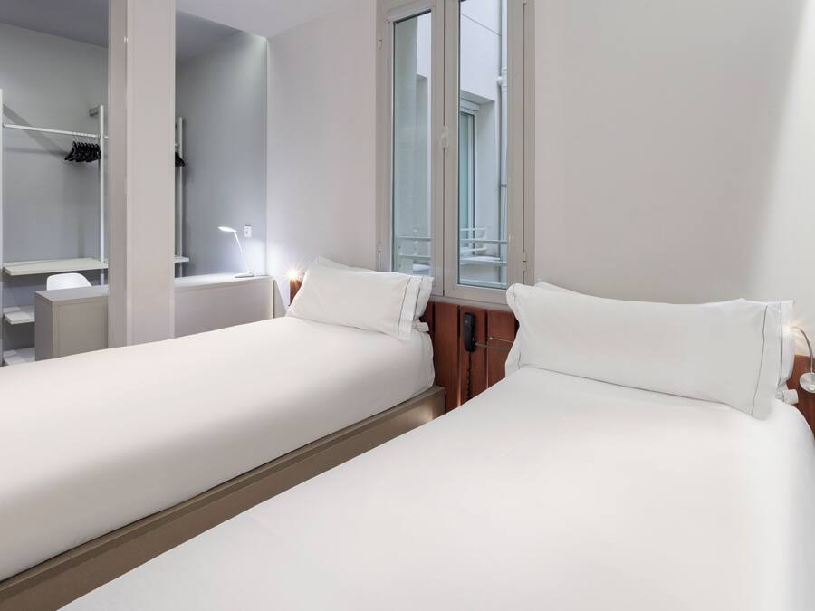 Une chambre d’hôtel avec deux lits simples, une table de chevet avec une lampe, un miroir, une fenêtre donnant sur l’extérieur et une armoire intégrée avec une tringle pour suspendre les vêtements.