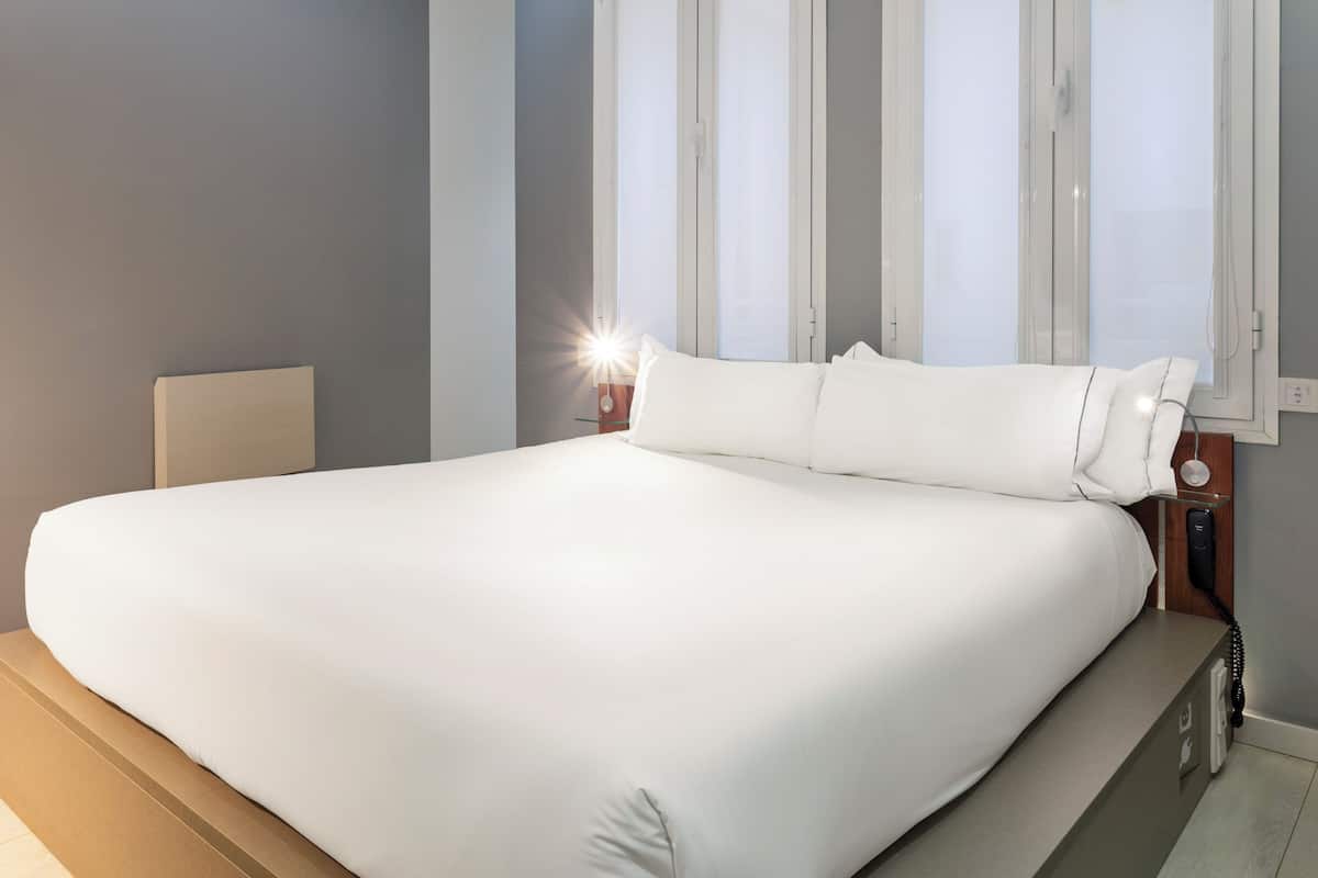A large bed with white linens, a bedside table with a lamp, and a window with blinds.