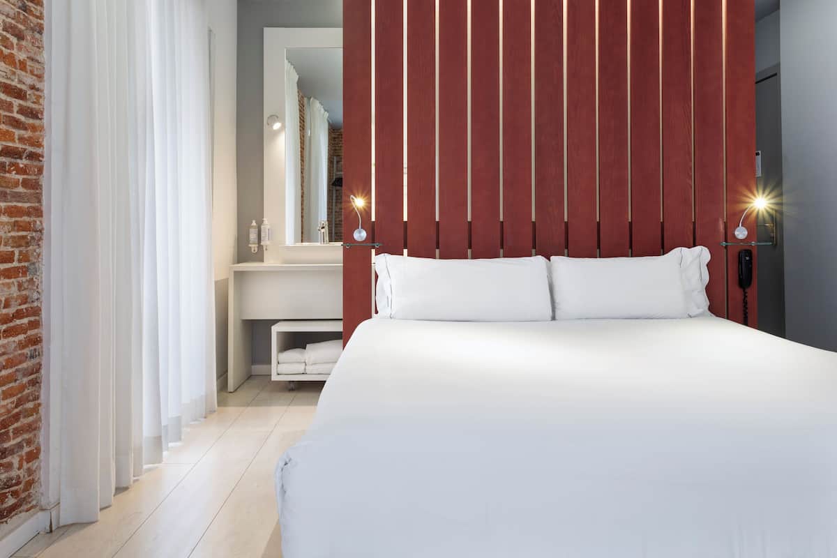 A modern hotel room with a large bed, a wooden panel wall, a bathroom with a mirror, and a brick wall.