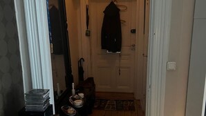 Un couloir avec un plancher en bois, une porte blanche et un manteau accroché au mur. On y voit une petite table sur laquelle sont posés quelques objets, et un téléviseur est visible au premier plan.