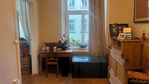 Une pièce étroite avec un bureau en bois, une chaise et un radiateur. Une fenêtre avec des rideaux laisse entrer la lumière naturelle. On peut voir un coffre noir et une armoire en bois contenant des photos encadrées.