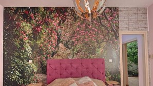 A bedroom with a tufted headboard, a large bed with patterned bedding, a wall mural of a garden, and a hanging spherical light fixture. - Ruby Apartment (Αγιος Νικολαος)