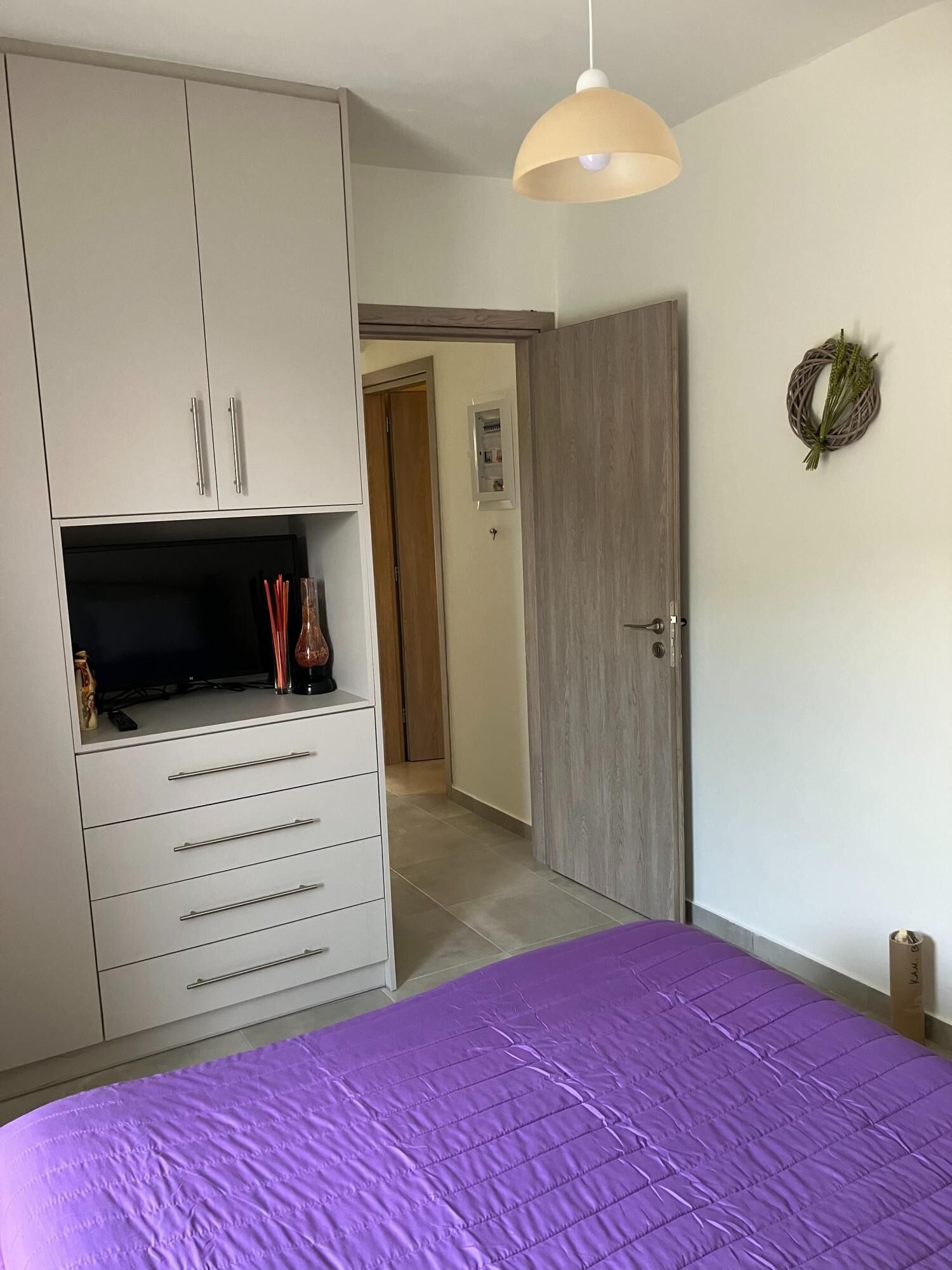 Quarto com colcha roxa, guarda-roupa branco com gavetas, uma televisão em cima e uma porta que dá para outro cômodo.