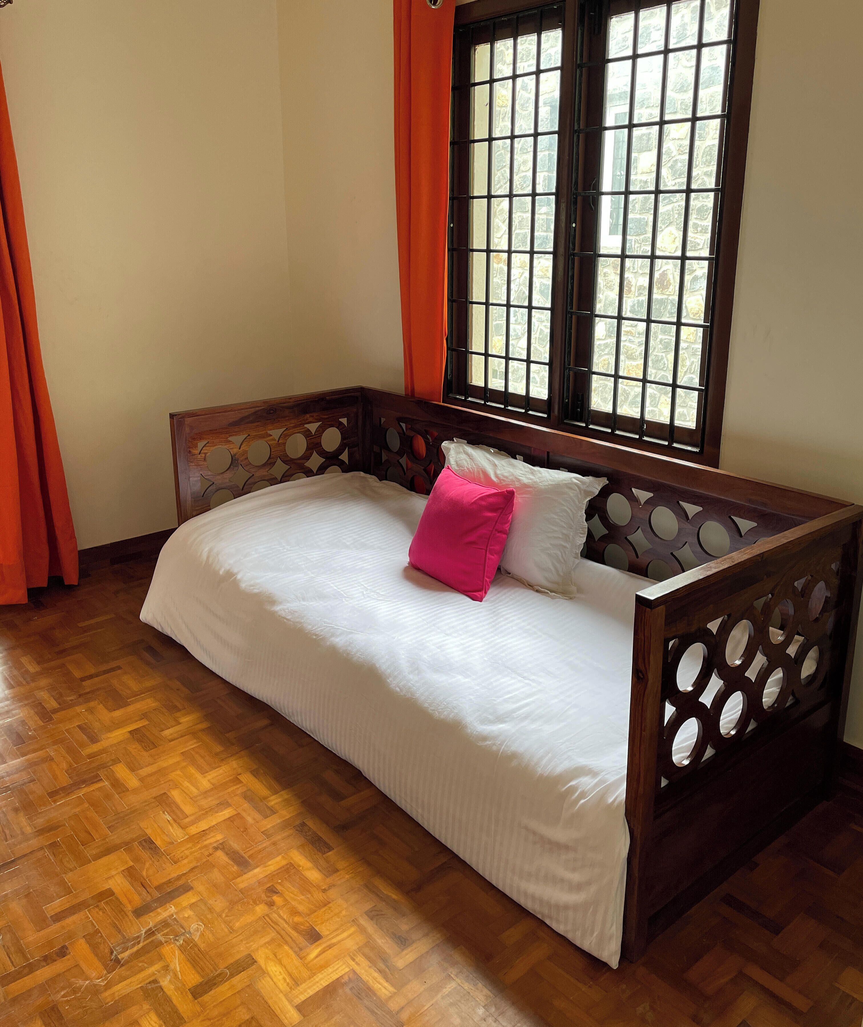 A wooden bed with a white mattress and two pillows, one pink and one white, in a room with a herringbone floor pattern and a window with a view of a stone wall.