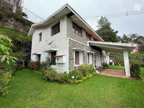 Una casa de dos pisos con fachada de piedra, un porche cubierto y un jardín bien cuidado con diversas plantas y flores.