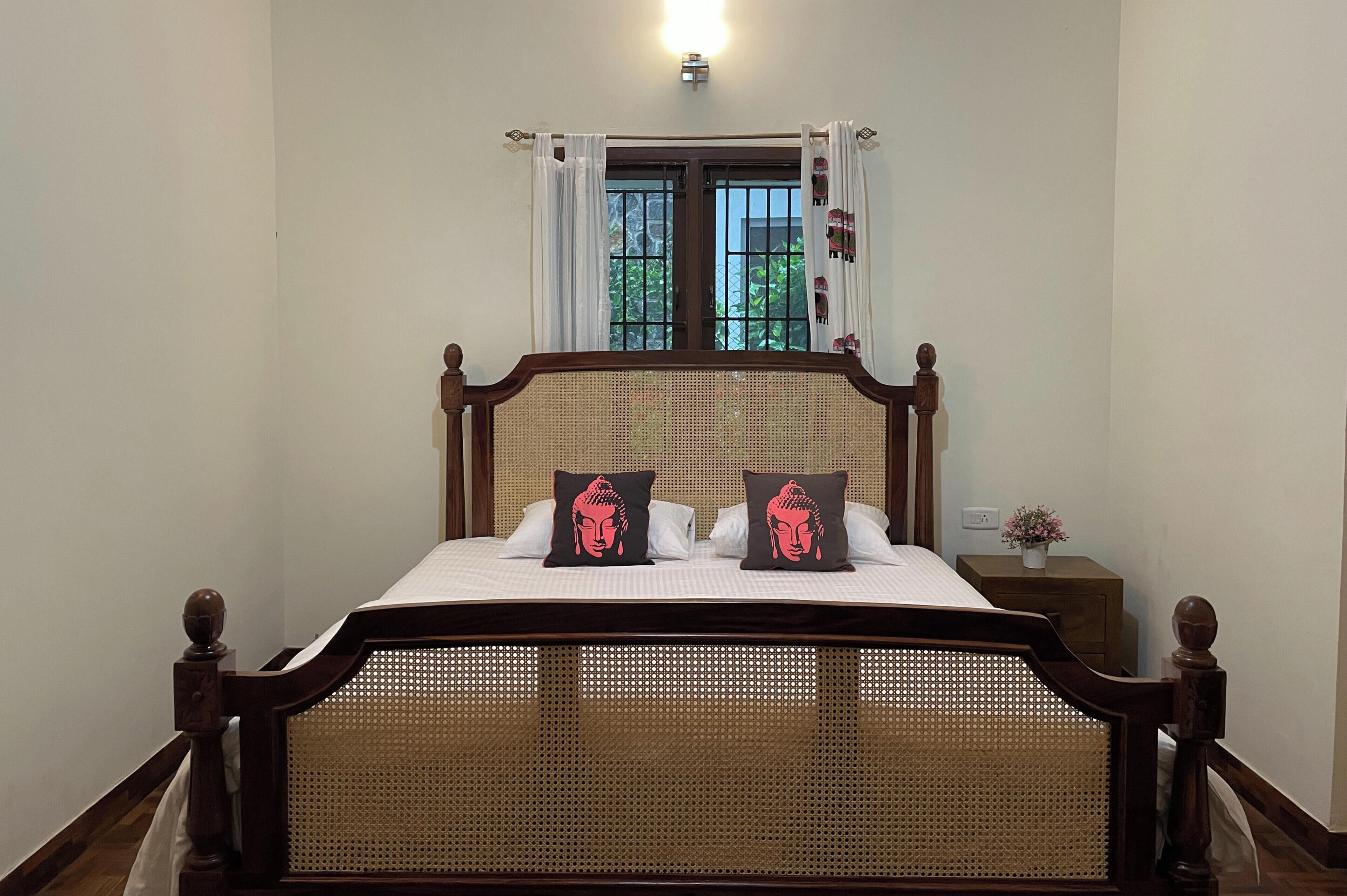 A bed with a wooden frame and a woven headboard, two pillows with a Buddha design, a nightstand with a small plant, and a window with a metal grill and curtains.