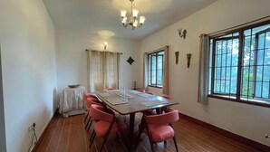 A dining room with a wooden table, red chairs, a chandelier, and large windows. - Nashika Villa by evaddo stays (Kodaikanal)
