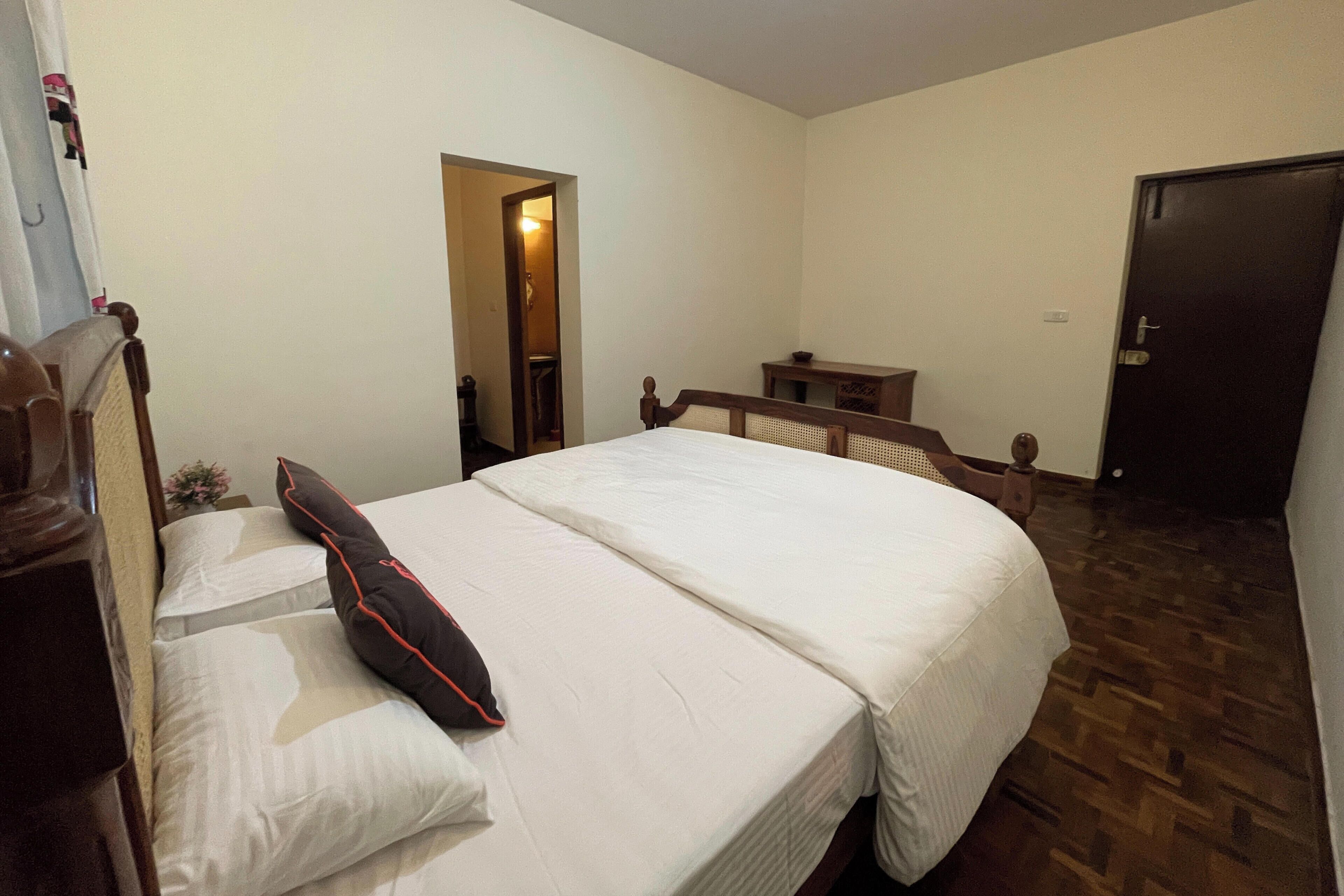 A bedroom with a large bed, a wooden nightstand, a desk, and a door.