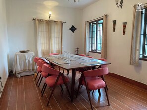 A dining area with a long table, red chairs, a small table with a bowl, and a window with curtains. - Nashika Villa by evaddo stays (Kodaikanal)