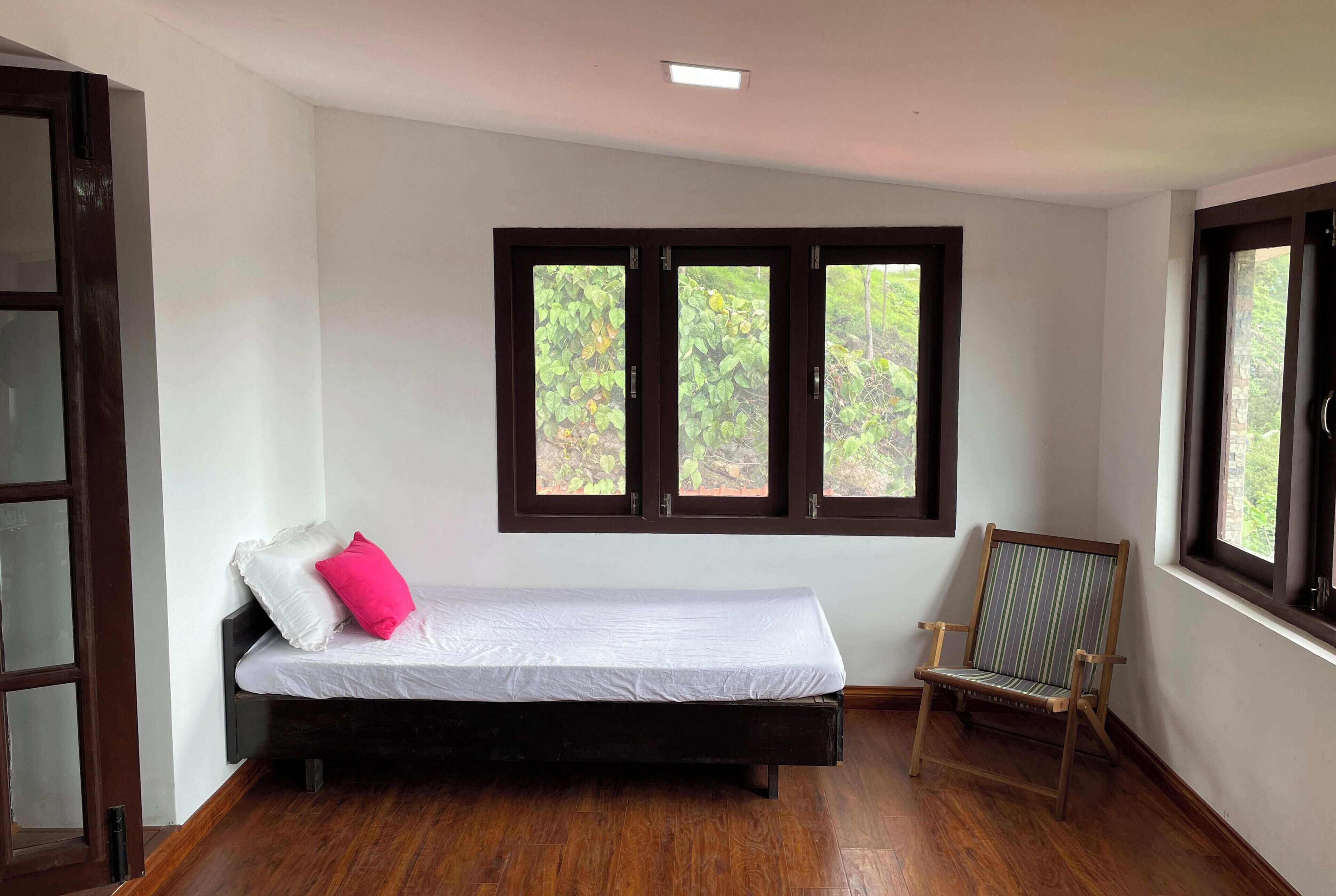 A room with a bed, a chair, and two windows with a view of greenery.