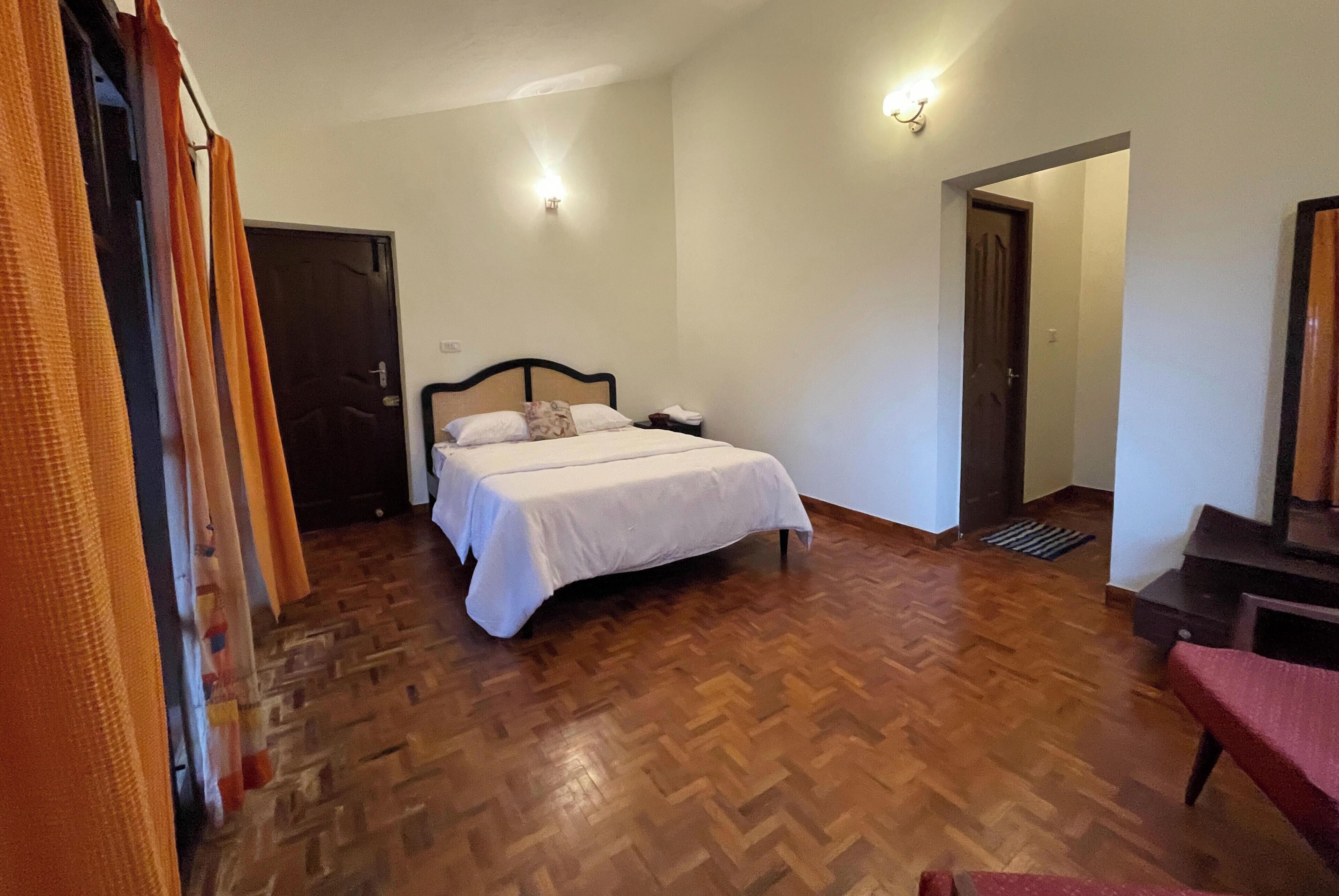 A bedroom with a wooden floor, a bed with white bedding, a nightstand, and a wall-mounted light fixture.