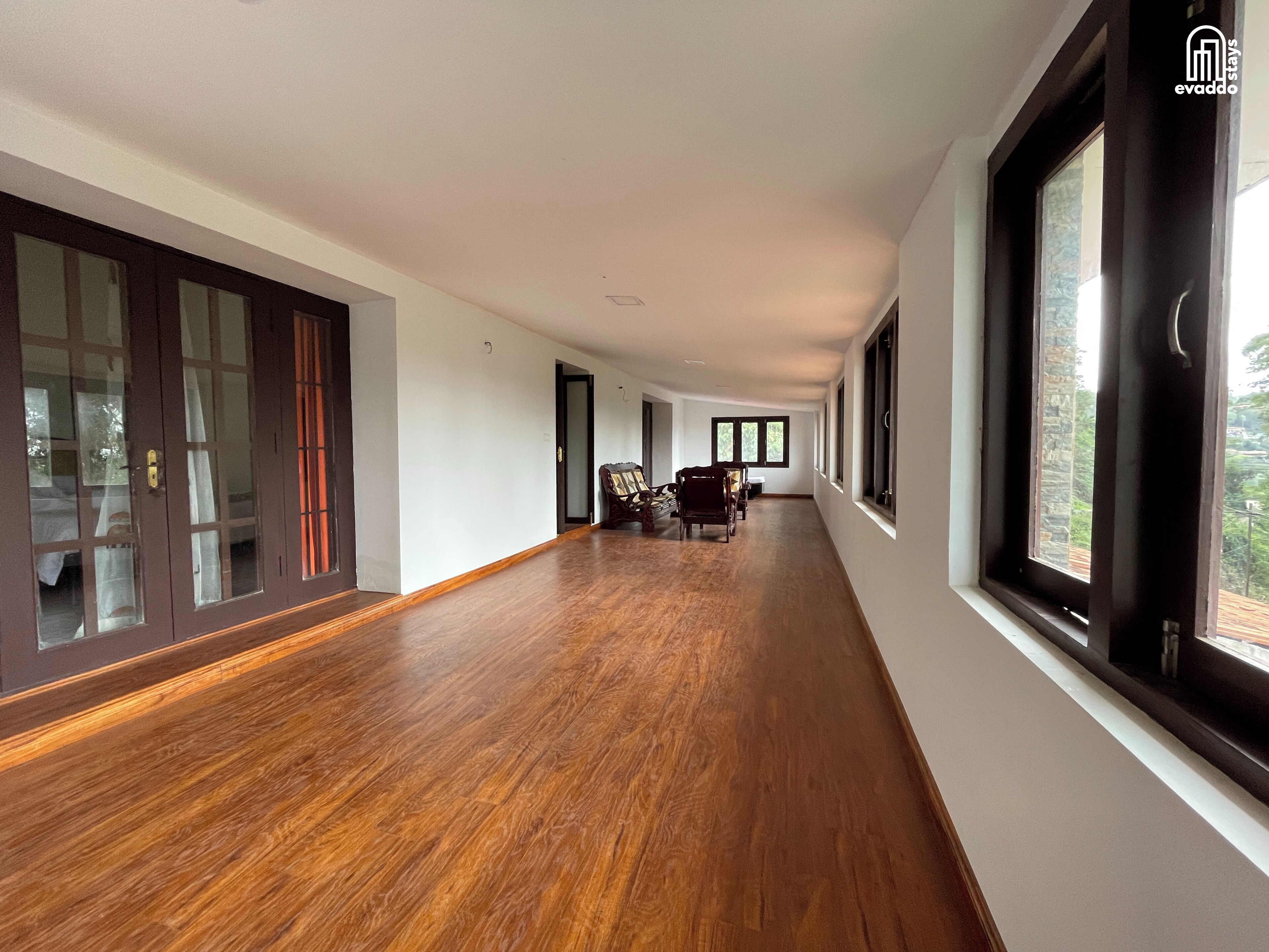 A long, open hallway with wooden flooring, large windows, and a dining area with chairs and a table.