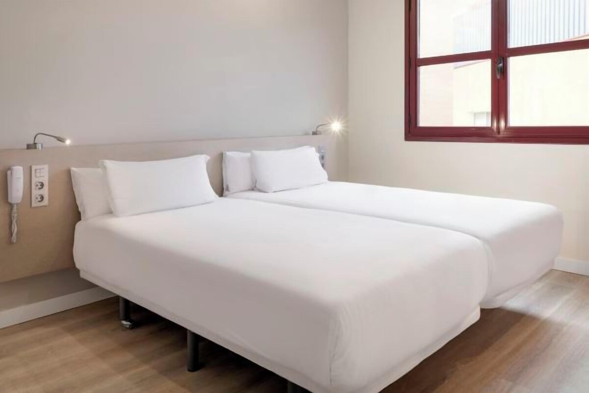A hotel room with two single beds, a wooden floor, a window with a red frame, and a wall-mounted phone.