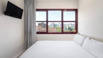 A hotel room with a large bed, a flat-screen TV mounted on the wall, and a window with a city view.