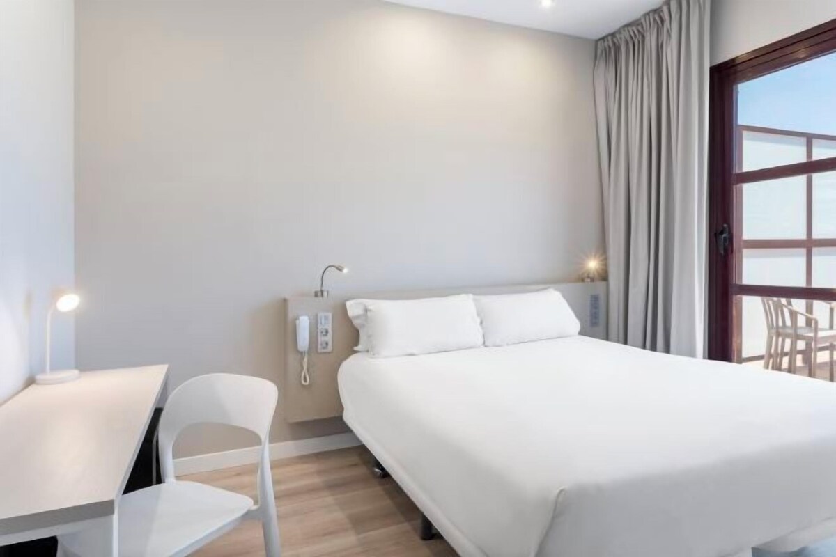 A hotel room with a bed, a desk, a chair, a phone, and a window with curtains.