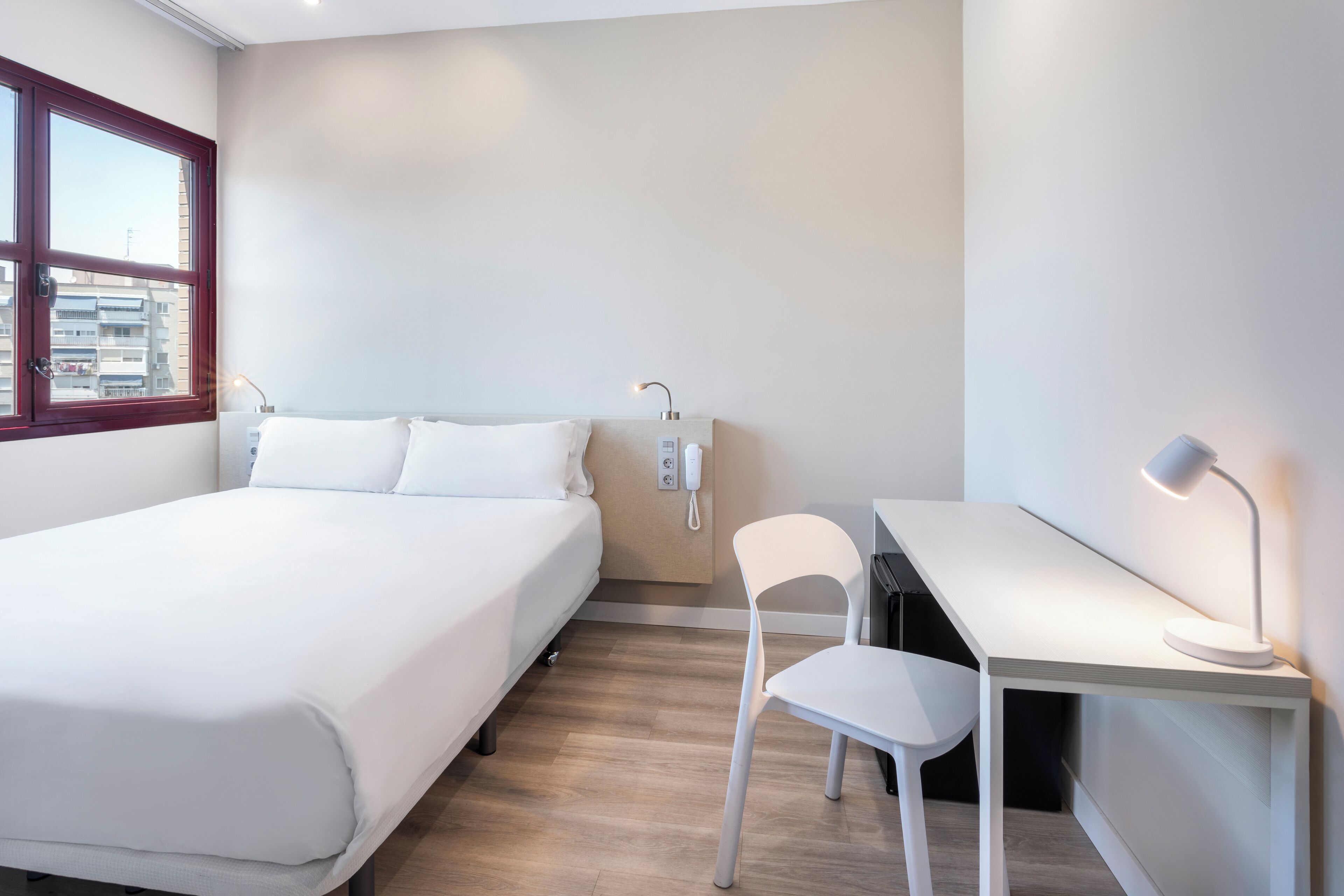 A modern hotel room with a bed, desk, chair, and a phone on the wall.