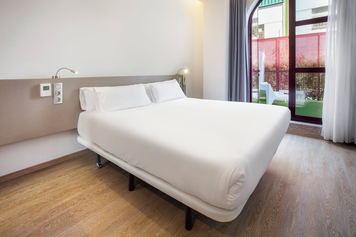 A neatly made bed in a hotel room with a wooden floor, a wall-mounted power outlet, and a window with a view of a red fence and greenery outside.