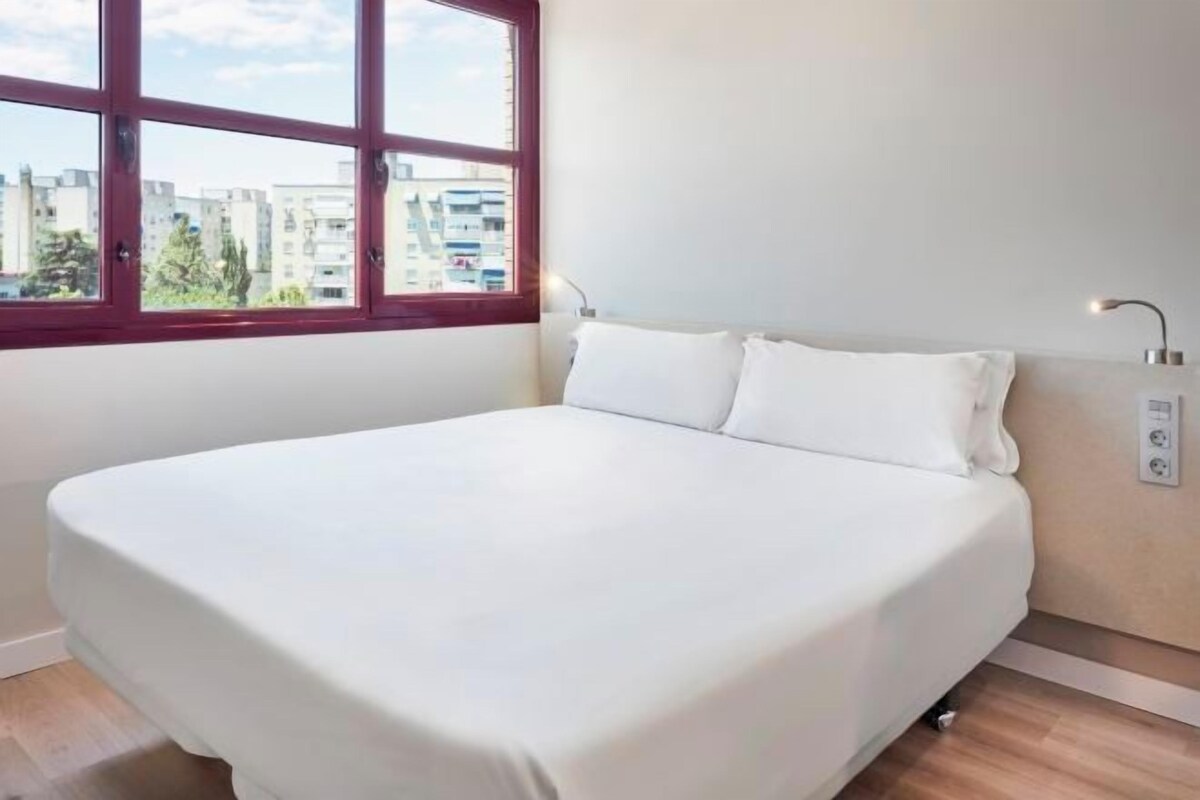 A neatly made bed in a room with a large window, a bedside table with a lamp, and a visible electrical outlet on the wall.