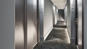 Un couloir d’hôtel moderne avec des portes numérotées de 005 à 006, un sol recouvert de moquette et un plafond en béton.