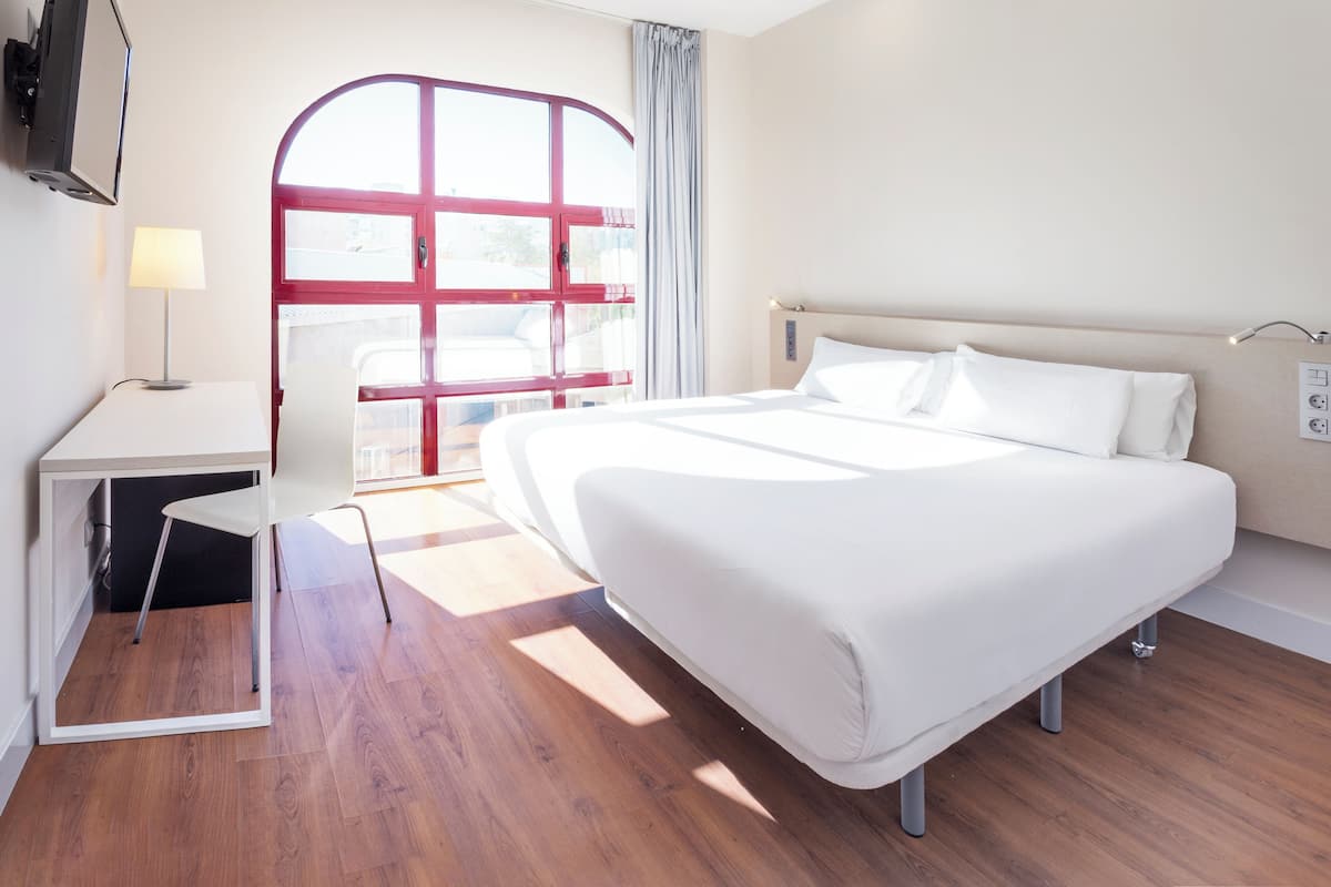 A modern hotel room with a large bed, a desk with a lamp, a TV, and a window with curtains.