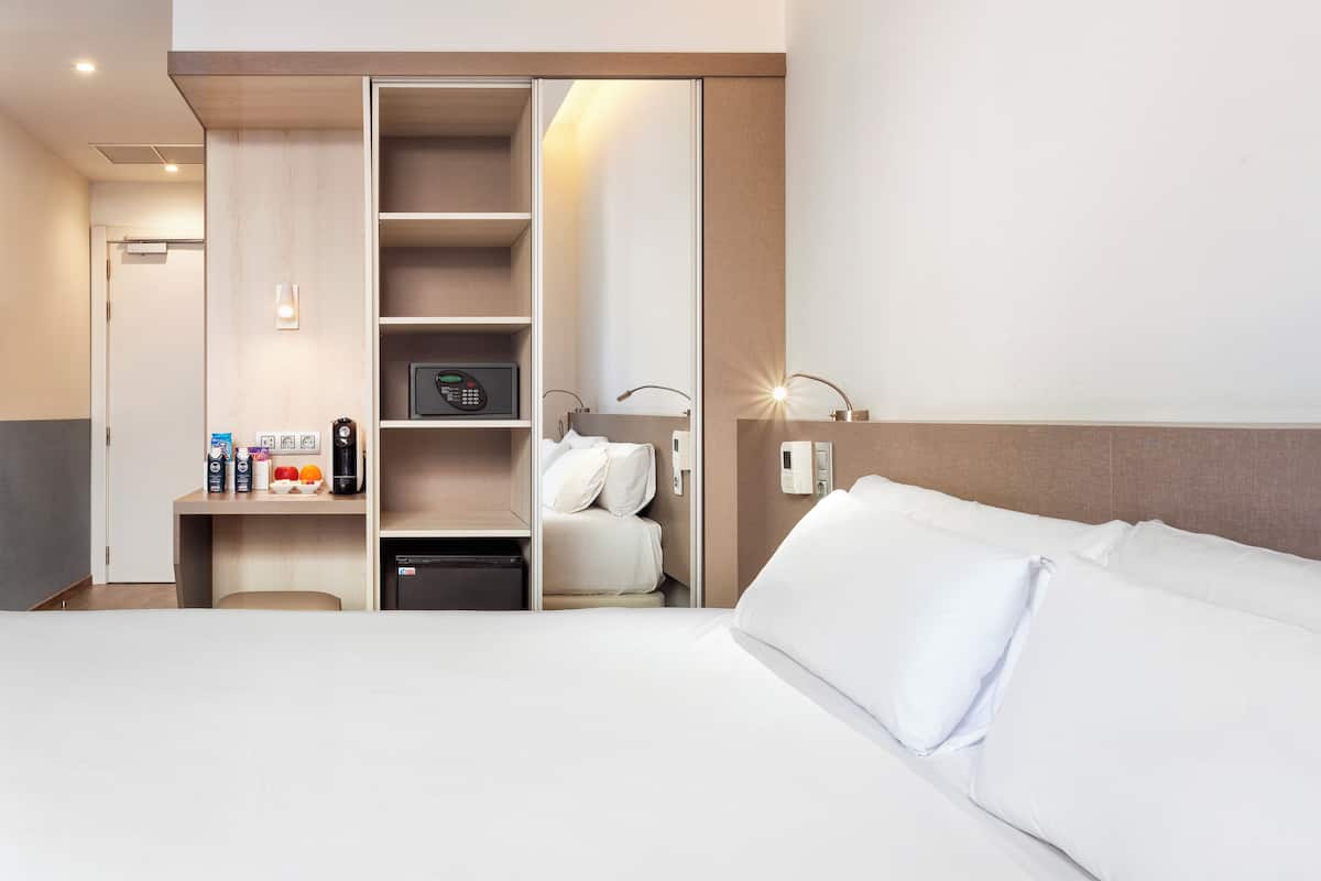 A hotel room with a bed, a desk with a coffee maker, a mini-fridge, a built-in wardrobe, and a bedside table with a lamp.