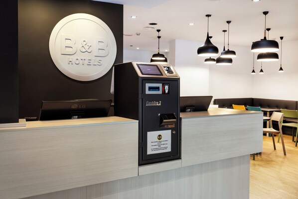 A hotel reception desk with a cash machine, a digital display, and a sign for B&B Hotels. - B&B Hotel Madrid Fuenlabrada (Fuenlabrada)