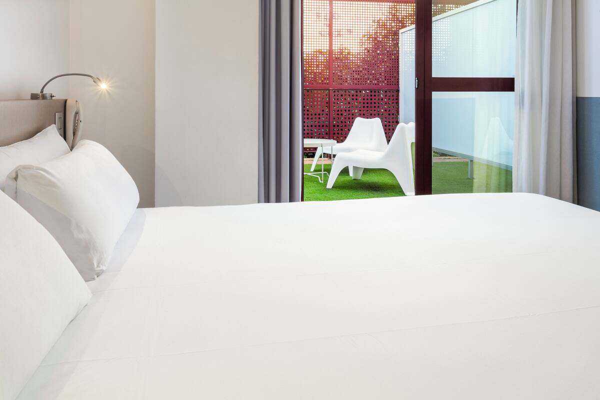 A modern hotel room with a large bed, a desk, and a view of a red fence and green lawn outside.