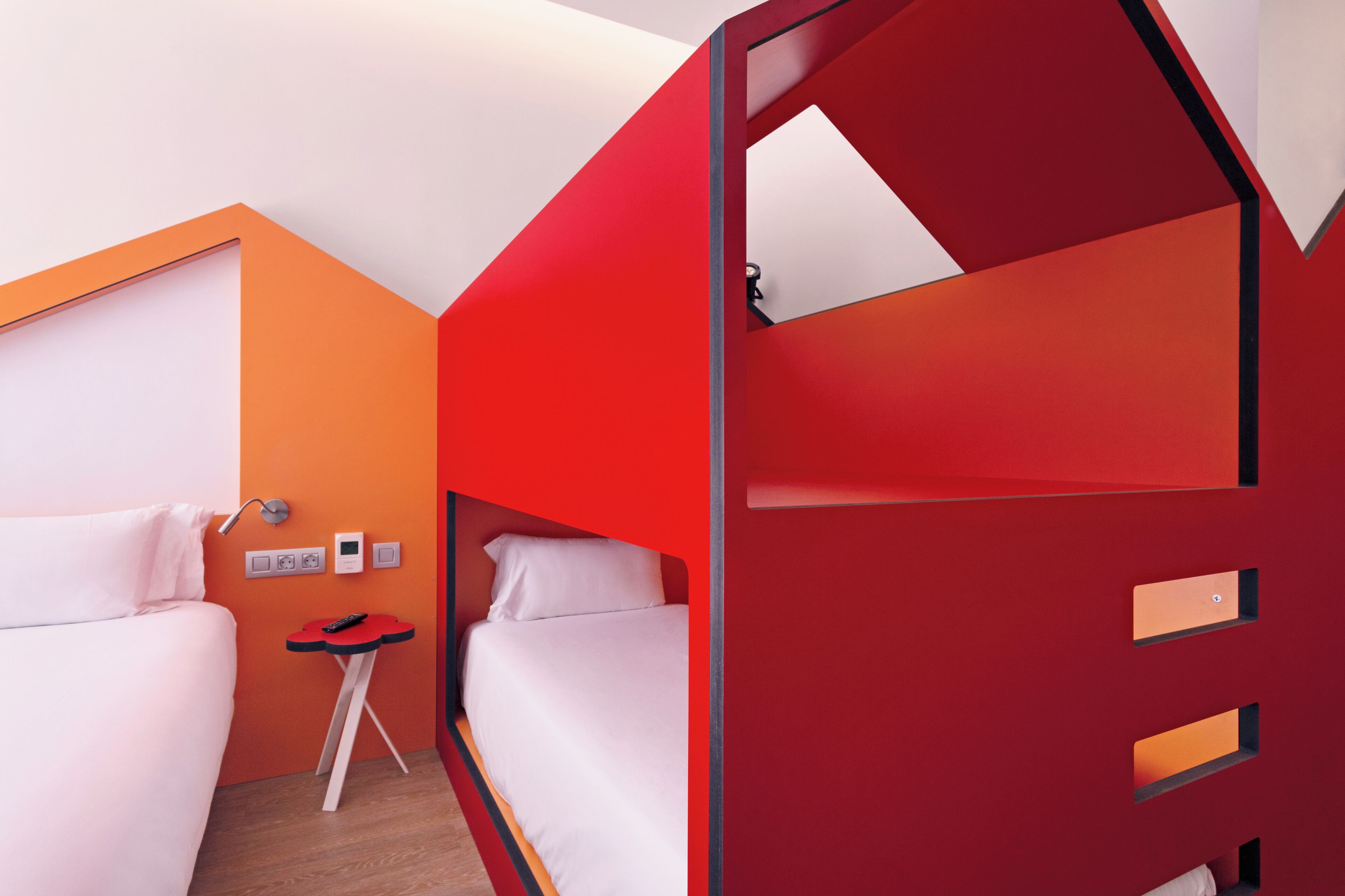 Una habitación de hotel moderna con una cama doble roja, una cama individual y una pequeña mesa con un control remoto.