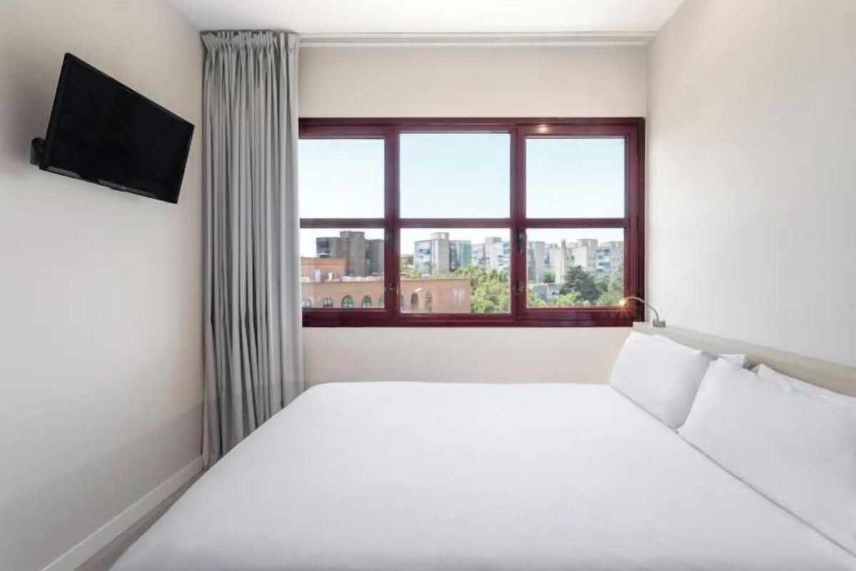 A hotel room with a large bed, a flat-screen TV mounted on the wall, and a window with a view of buildings.