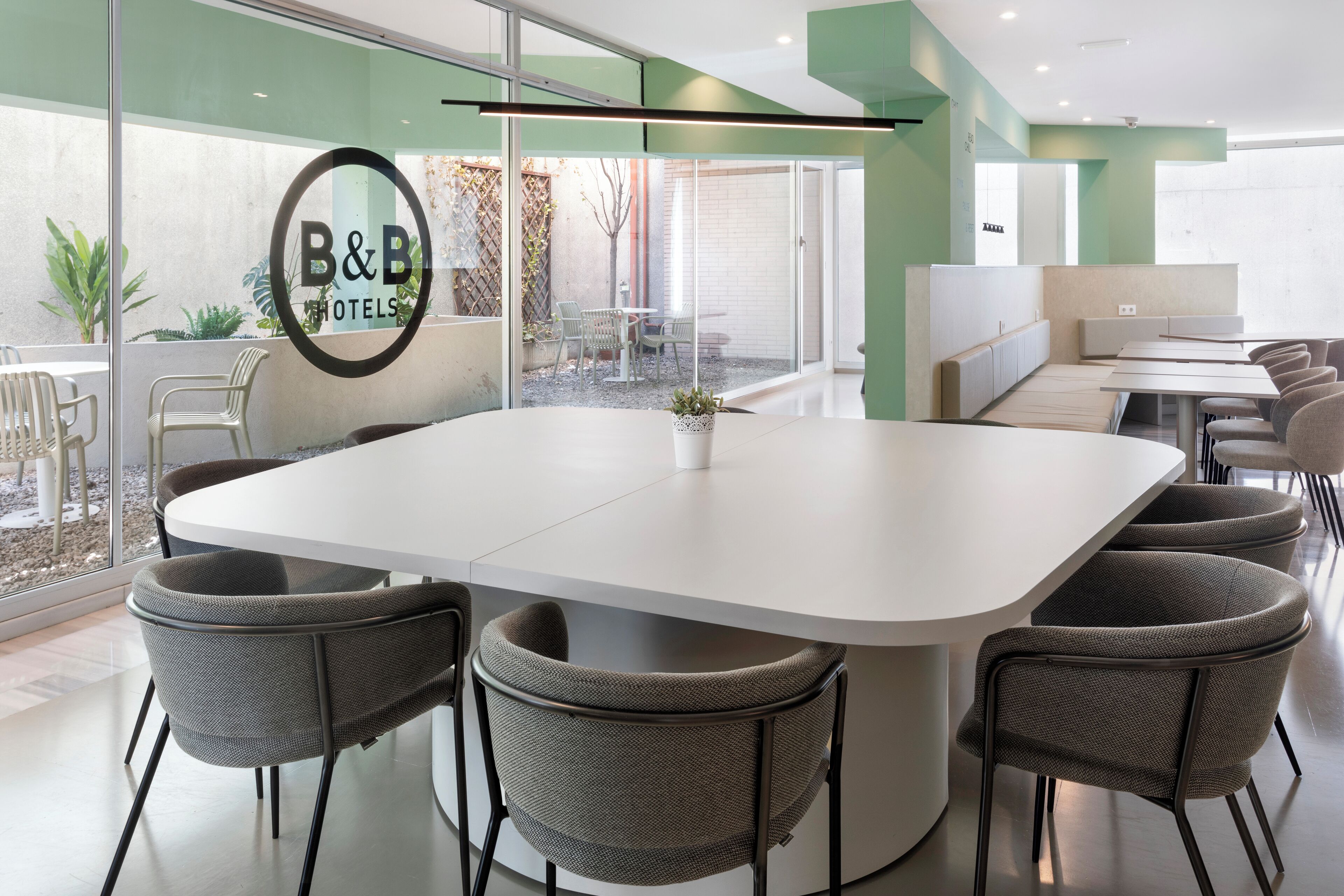 A modern hotel conference room with a large table, comfortable chairs, and a glass wall offering a view of the outdoor patio area.