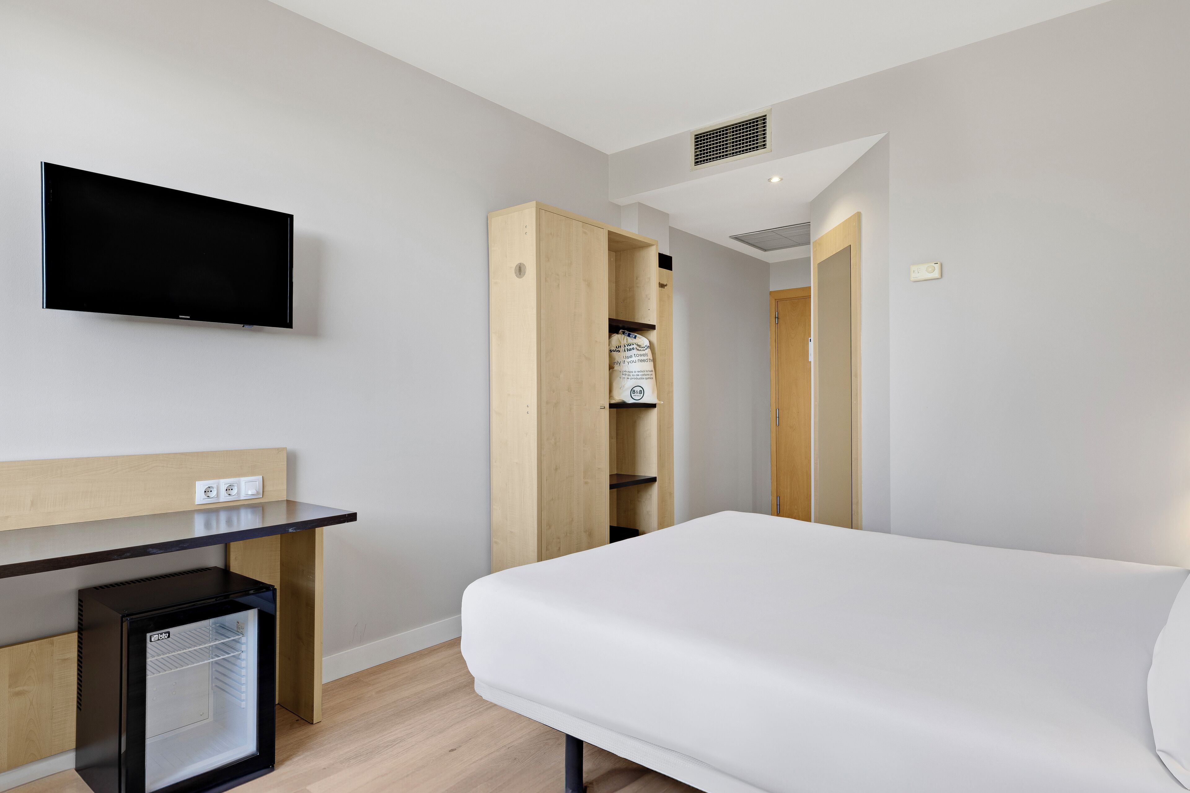 A hotel room with a bed, a TV mounted on the wall, a desk with a mini-fridge, and a wooden wardrobe.