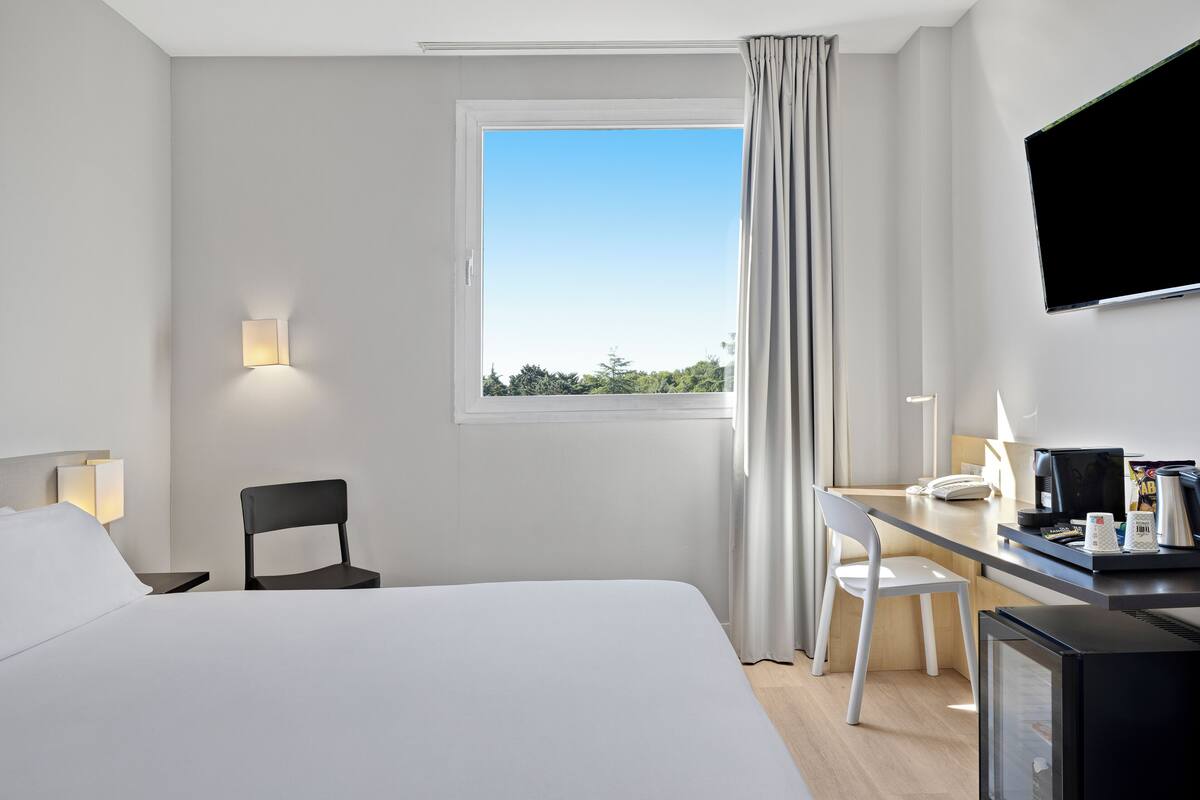 Ein Hotelzimmer mit einem Bett, einem Schreibtisch mit Computer, einem Sessel, einem Fernseher und einem Fenster mit Vorhängen.
