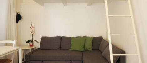 Un espacio habitable compacto con un sofá, una mesita, una silla y una escalera apoyada en la pared.