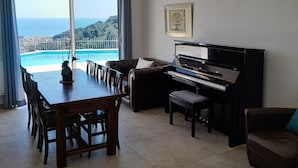 Un vaste salon comprenant un piano, une table à manger avec des chaises, un canapé et une grande fenêtre offrant une vue sur la mer et le paysage urbain.