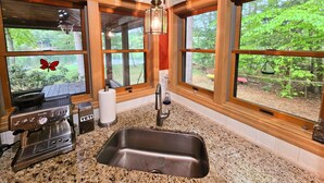 Una cocina con cafetera, un lavamanos y ventanas amplias con vista a un jardín.