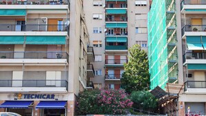 A multi-story residential building with balconies, a visible storefront with a sign reading "TECNICAR," and a tree with pink flowers in the foreground. - Golden skyview from San Martino by Wonderful Italy (Genoa)