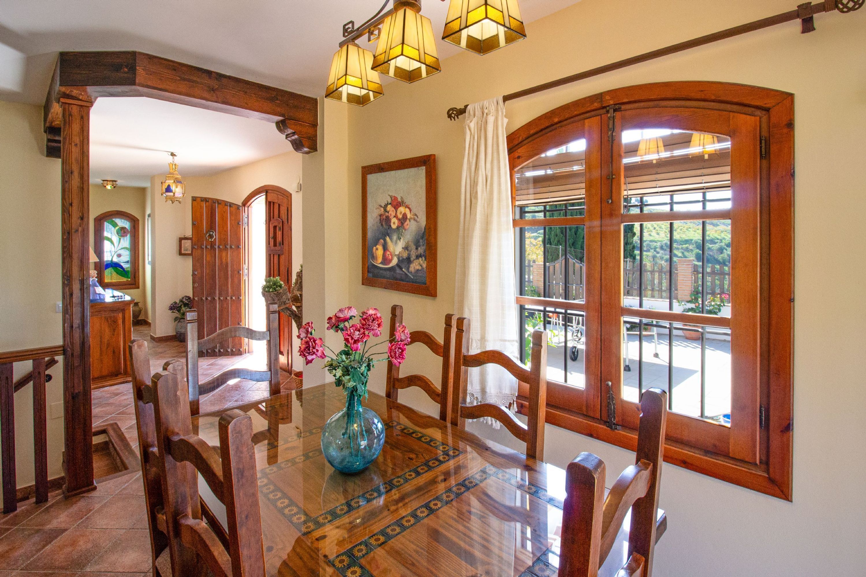 Un comedor con una mesa de madera, sillas y un jarrón de flores. Hay un cuadro en la pared y una ventana con cortinas.