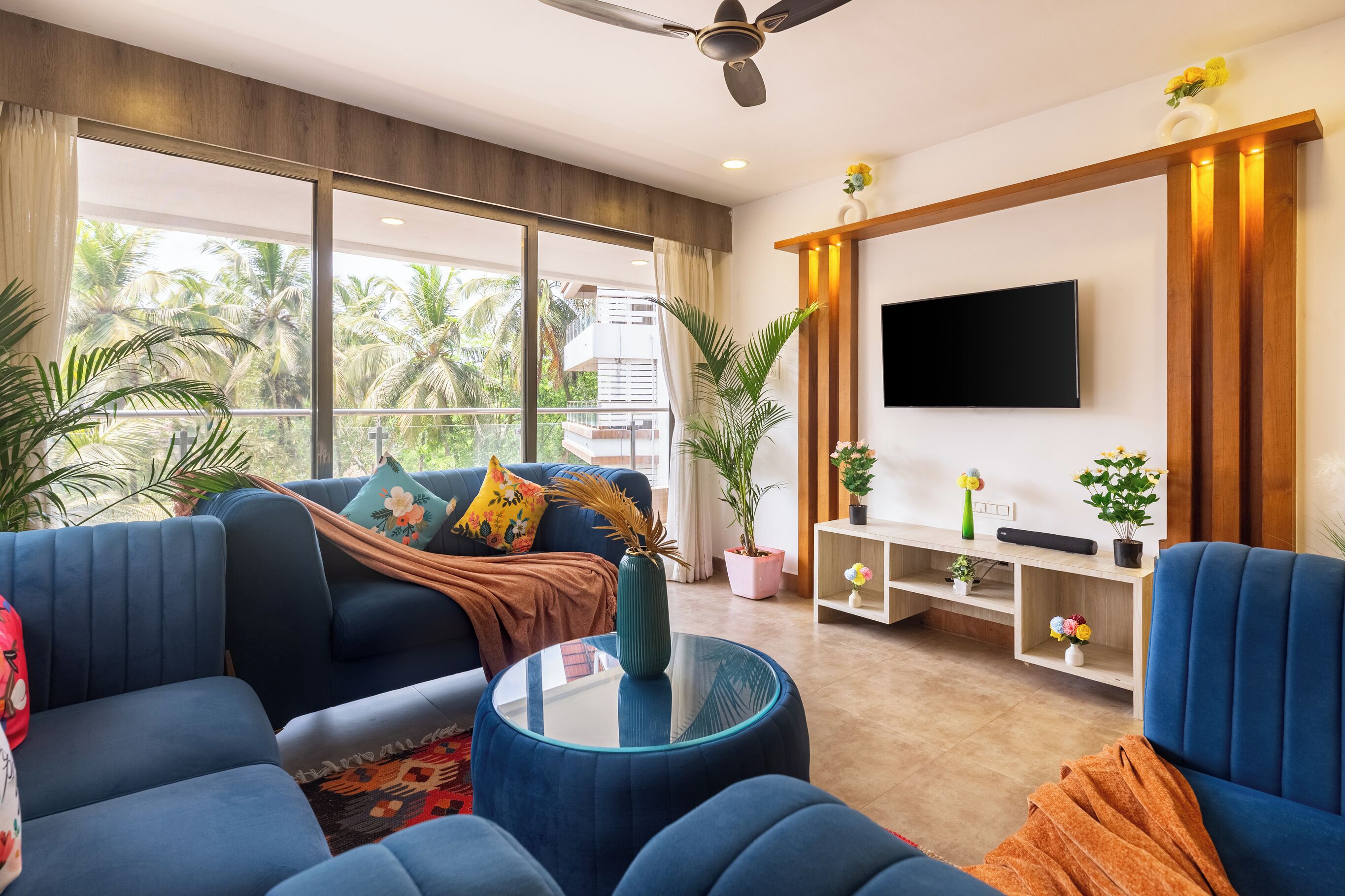 A modern living room with a sofa set, a coffee table, a TV, and large windows with a view of palm trees.