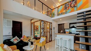Una sala moderna con un sofá, un sillón amarillo y una pequeña mesa redonda. Hay una zona de cocina con una encimera y taburetes.