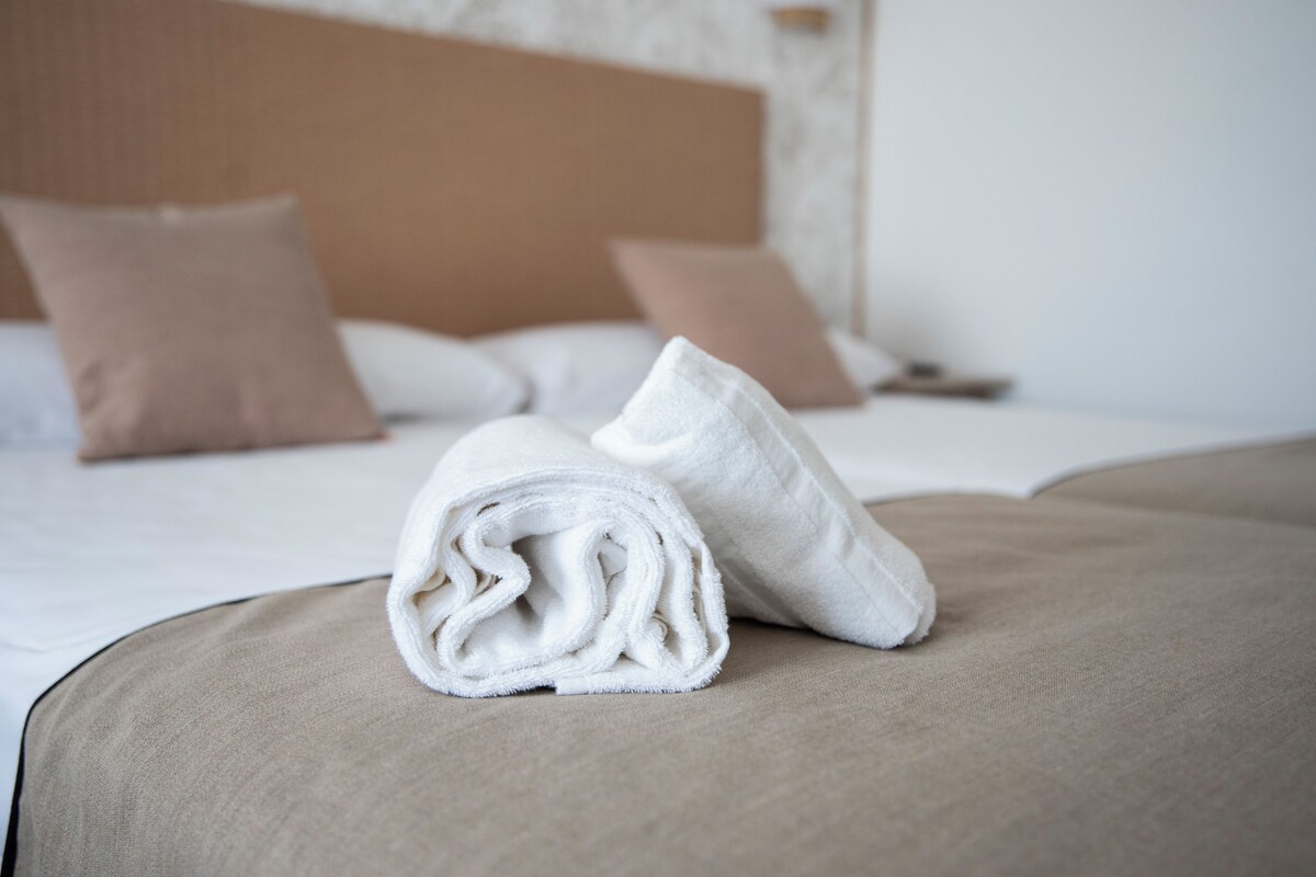 A neatly made bed with a beige bedspread and pillows, a rolled white towel on the bed, and a headboard with a wooden panel.