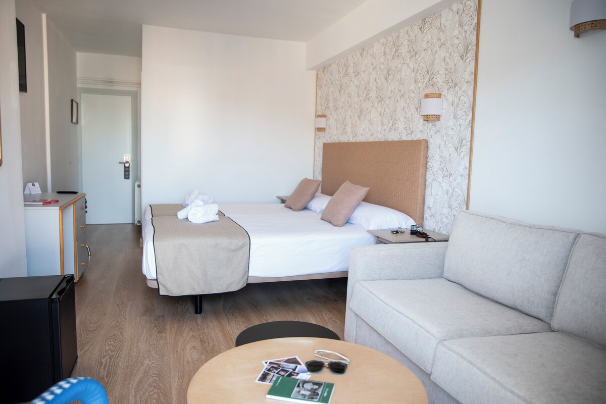 A hotel room with a bed, a sofa, a small table with items, and a desk with a computer.